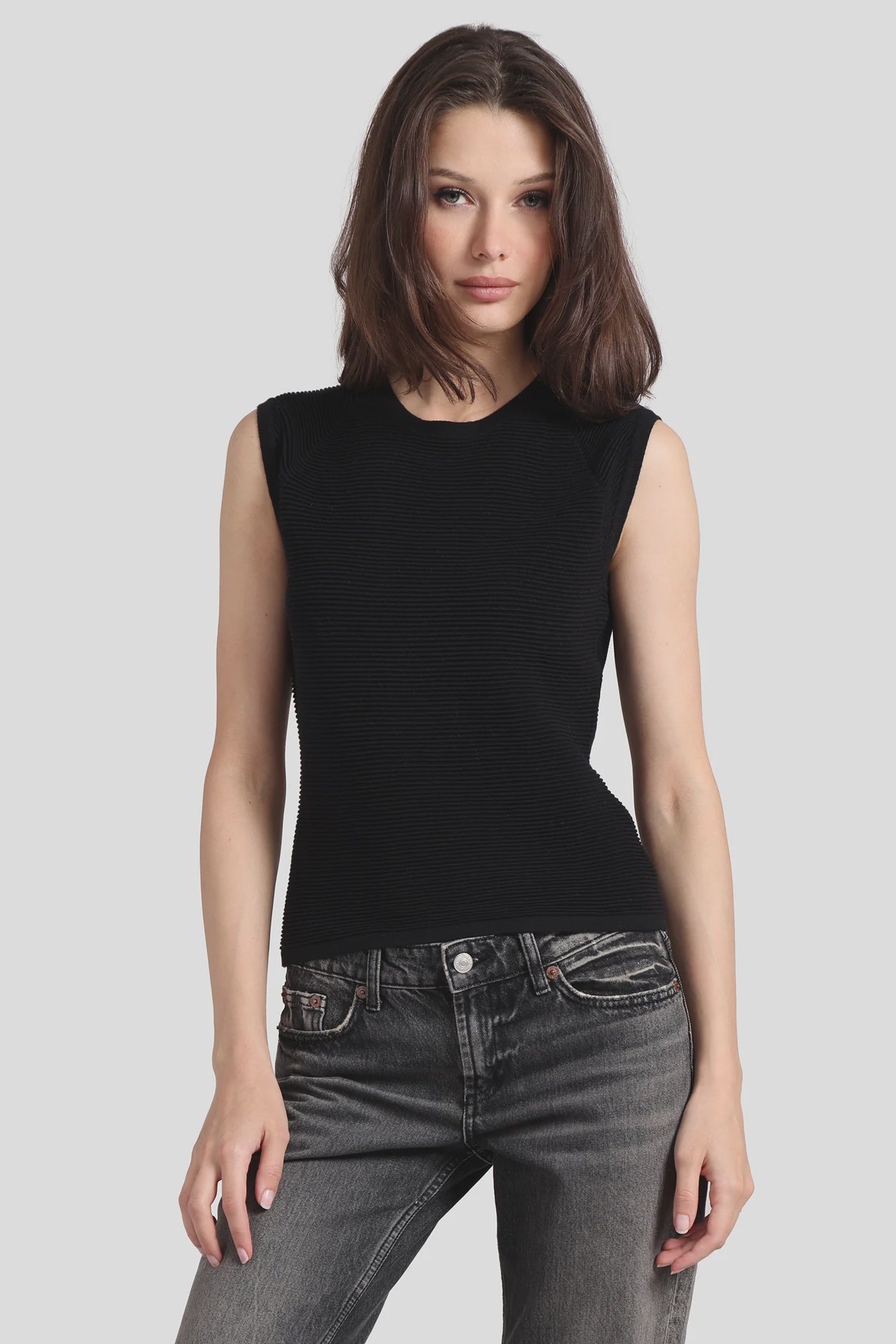 A woman with shoulder-length brown hair is wearing a sleek Ottoman Sleeveless Top by Minnie Rose. The black top features intricate rib detailing and is paired with dark jeans. She stands against a plain light gray background, looking directly at the camera.