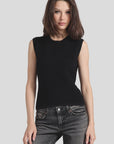 A woman with shoulder-length brown hair is wearing a sleek Ottoman Sleeveless Top by Minnie Rose. The black top features intricate rib detailing and is paired with dark jeans. She stands against a plain light gray background, looking directly at the camera.