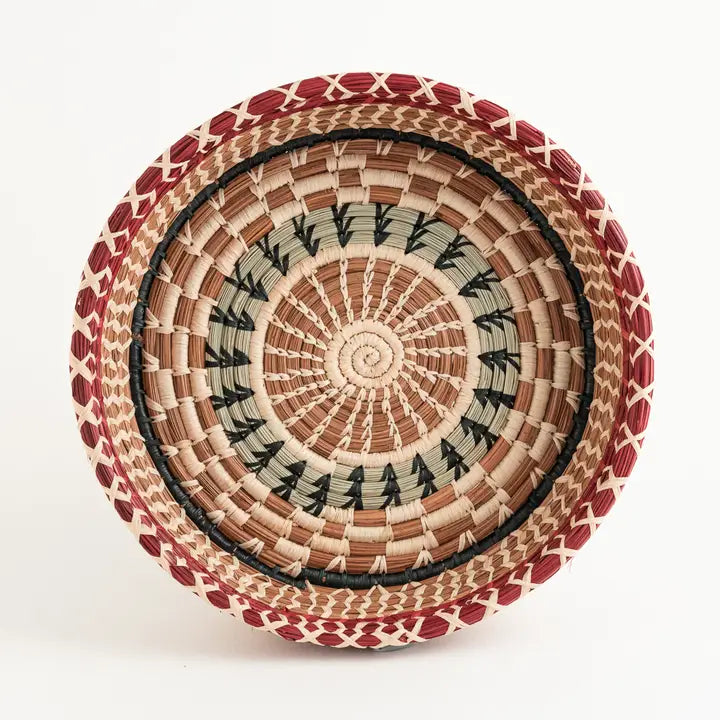 The Faire Manuela Basket is a round Guatemalan basket with intricate patterns in natural, tan, black, and red, featuring hand-dyed raffia highlights and geometric accents on the rim and interior.