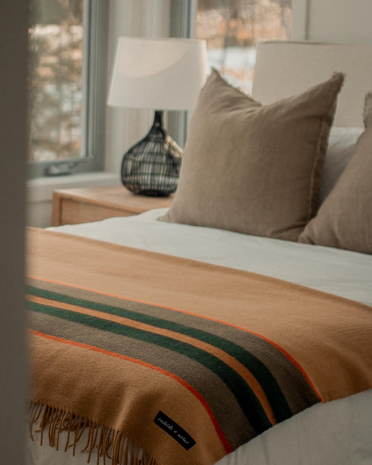 A cozy, sunlit bedroom features a neatly made bed with beige pillows, a vintage-inspired Faire S&A Blanket with green and orange stripes, and a bedside table topped by a black lamp.