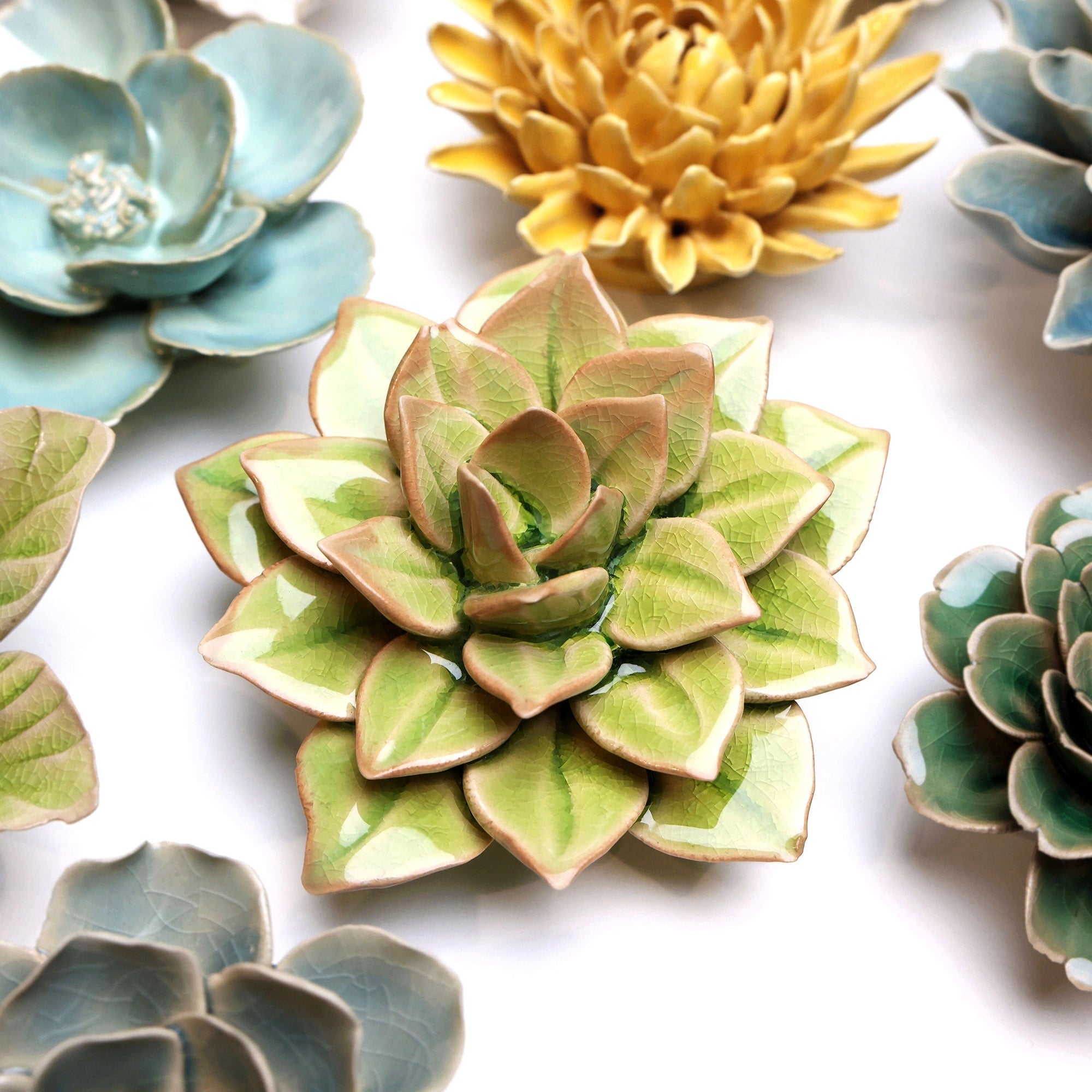 Chive Ceramics (thru Faire) Green Fairy Echeveria is a coastal-inspired ceramic succulent, showcased with green, yellow, and blue floral sculptures arranged on a white surface. The centerpiece features layered leaves resembling a real echeveria.