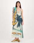 A woman with long brown hair wears the Me369 Olivia Camisole Dress featuring palm trees and "AMAZING THINGS WILL HAPPEN" text, styled with a patterned scarf and gold shoes against a plain background.