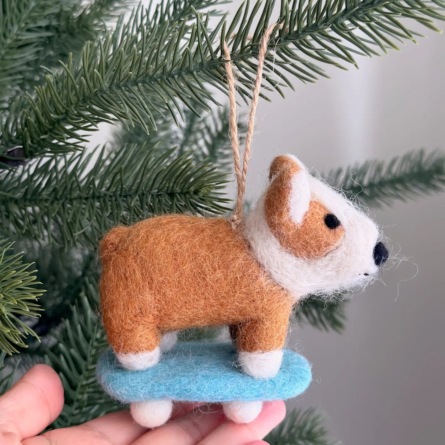 A hand displays the Faire Felted Ornament, a brown and white bulldog on a blue skateboard—an adorable, unique decoration for your Christmas tree.