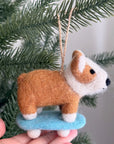 A hand displays the Faire Felted Ornament, a brown and white bulldog on a blue skateboard—an adorable, unique decoration for your Christmas tree.