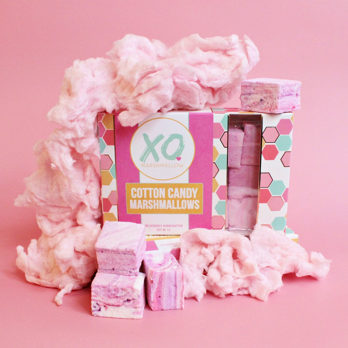 A box of Faire Cotton Candy Marshmallows, naturally colored and allergy friendly, rests on a pink surface with fluffy cotton candy and swirled pink-and-white marshmallows around it.