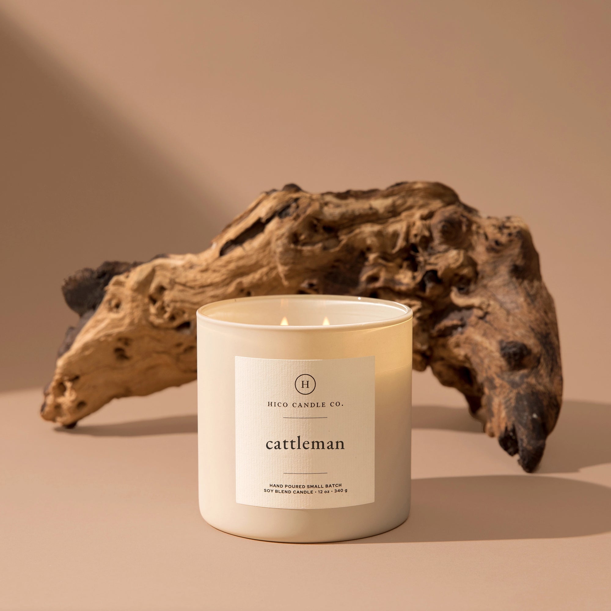 A white 14oz. "Cattleman" soy wax candle by Faire, known for its masculine scent, is placed in front of weathered driftwood on a beige surface with a neutral background.