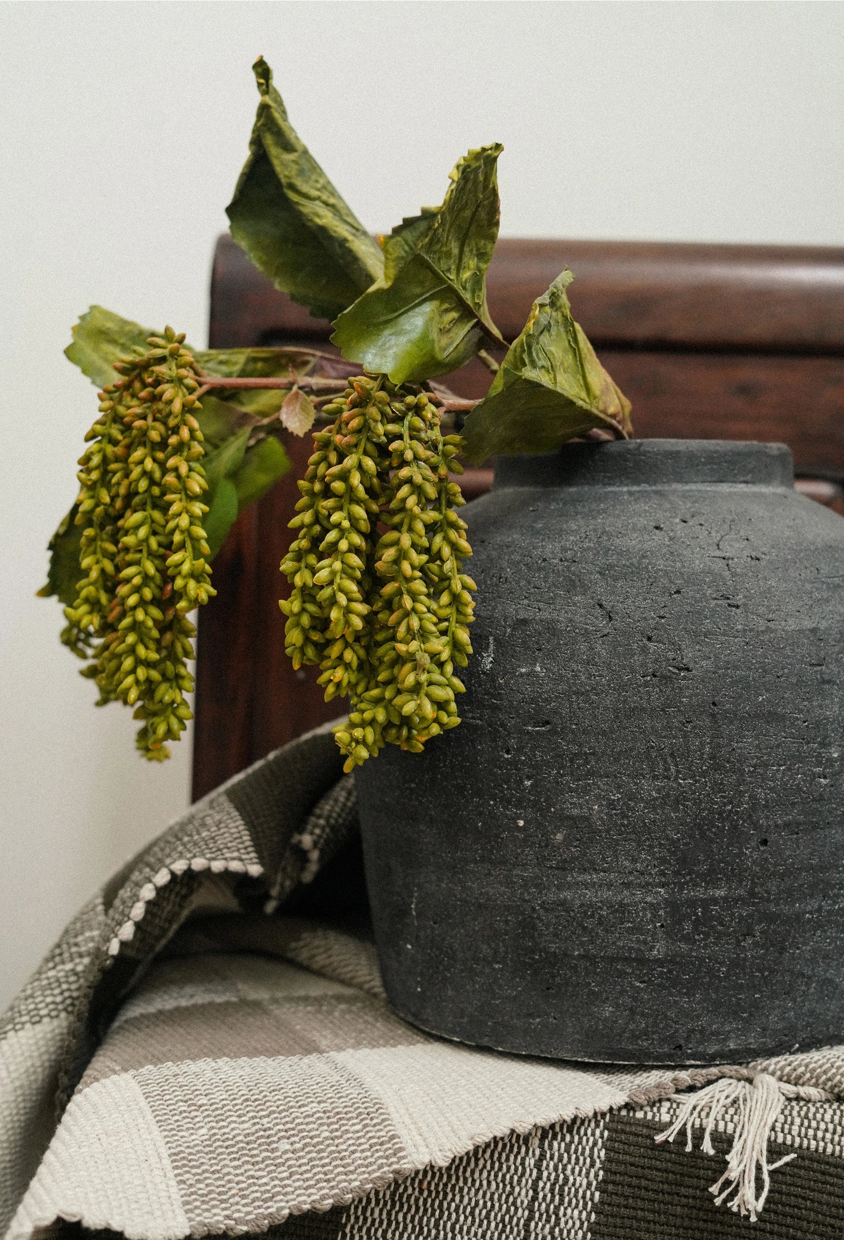 The Indaba Dripping Seed Stem, with clusters of green seed pods and dry leaves, cascades from a dark ceramic vase set on a striped neutral cloth against wood—an elegant botanical accent.