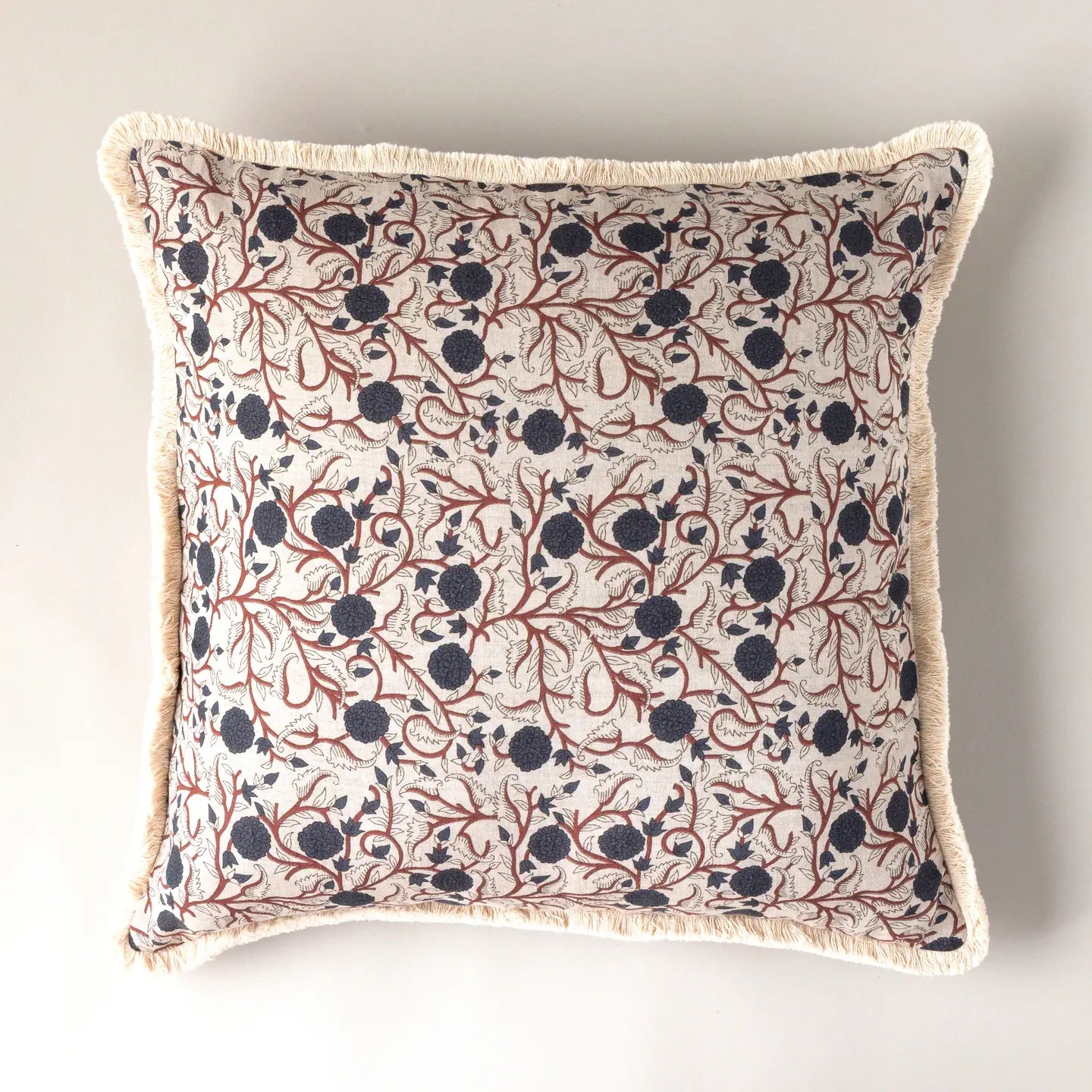 The Block Print Pillow by Faire features a detailed botanical print of dark blue berries and red branches on a cream background with off-white fringe trim. This vintage throw pillow is shown against a plain light surface.