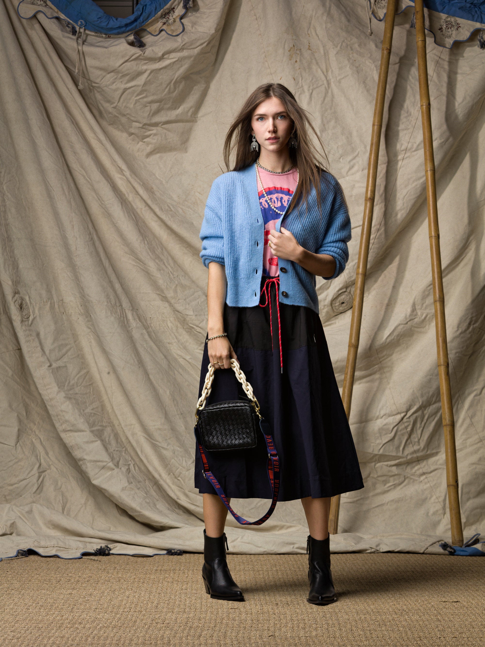 A woman stands before a beige draped backdrop, wearing a blue cardigan over a pink graphic tee, the Ping Pong Surf Club Le Vintage Sport Skirt, black ankle boots, and holding a black woven handbag with a chunky strap.