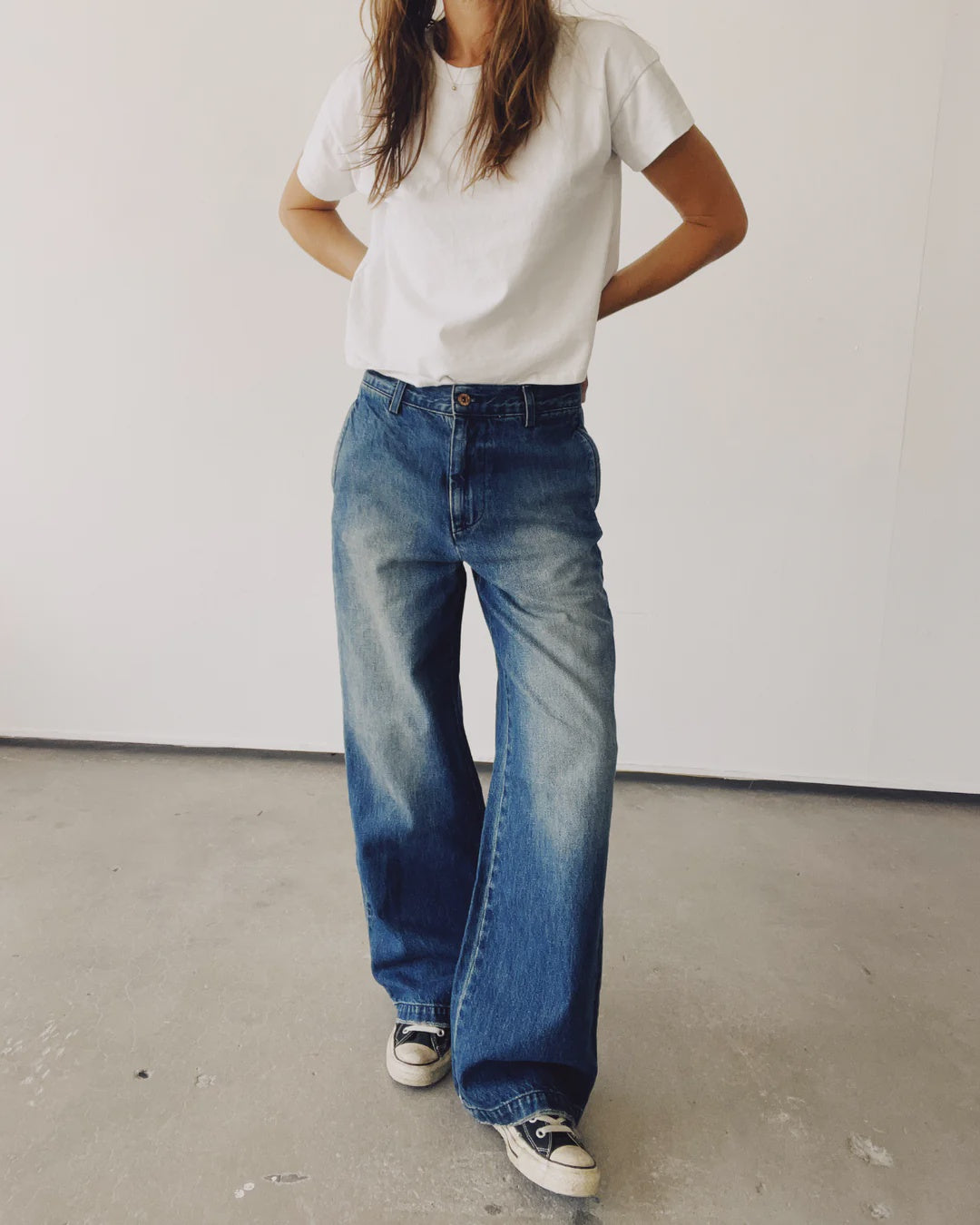 A person wearing a white t-shirt and The Buckle Back Trouser from Hey Gang, which are wide-leg, cotton denim jeans with a slouchy fit, stands on a concrete floor. They have black and white sneakers on and their hands on their hips against a plain white wall.