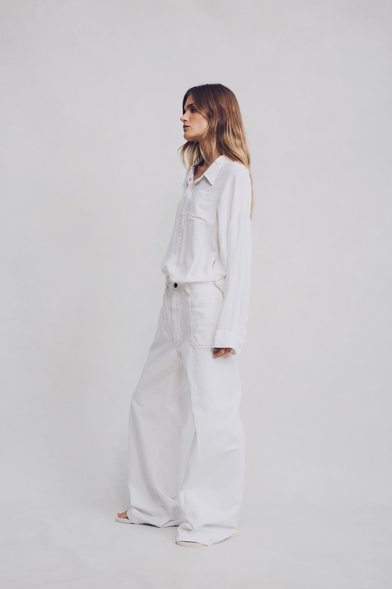 A woman stands against a plain light background, wearing a loose-fitting white button-up shirt and Denimist’s 100% cotton DECK PANT WHITE WASH. Her long hair is down, and she faces to the side with a relaxed posture.