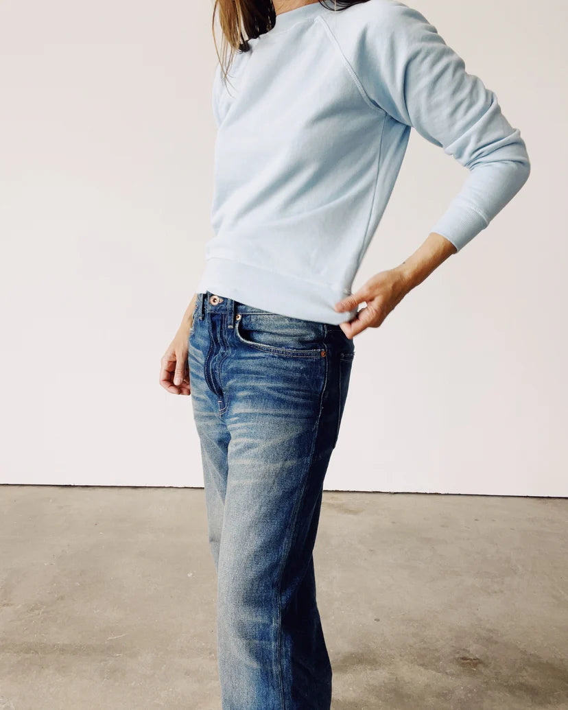 A person dressed in a vintage light blue &quot;The Sweatshirt&quot; by Hey Gang and faded blue jeans stands against a plain white wall. They have one hand tucked into their pocket while the other adjusts their sweatshirt, highlighting the casual ensemble.