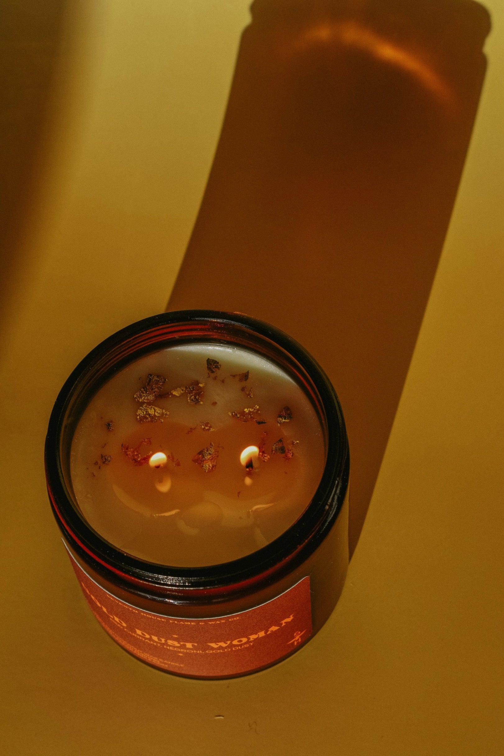 Brown jar Gold Dust Woman Candle by Faire with flower petals on a yellow surface, casting a long shadow.