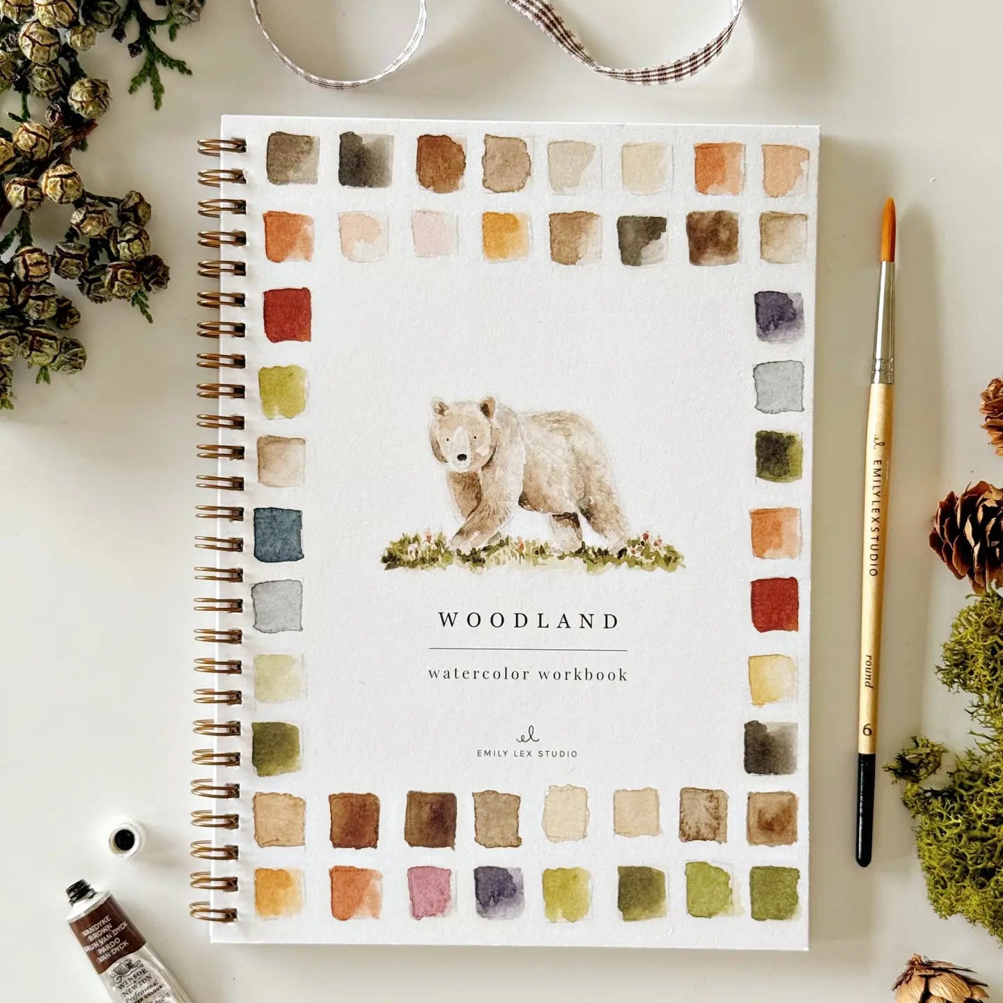 The Faire Watercolor Workbook is a spiral-bound, beginner-friendly guide with a bear cover illustration, watercolor swatches, and surrounded by a paintbrush, paint tube, pine cones, ribbon, and greenery.