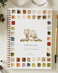 The Faire Watercolor Workbook is a spiral-bound, beginner-friendly guide with a bear cover illustration, watercolor swatches, and surrounded by a paintbrush, paint tube, pine cones, ribbon, and greenery.