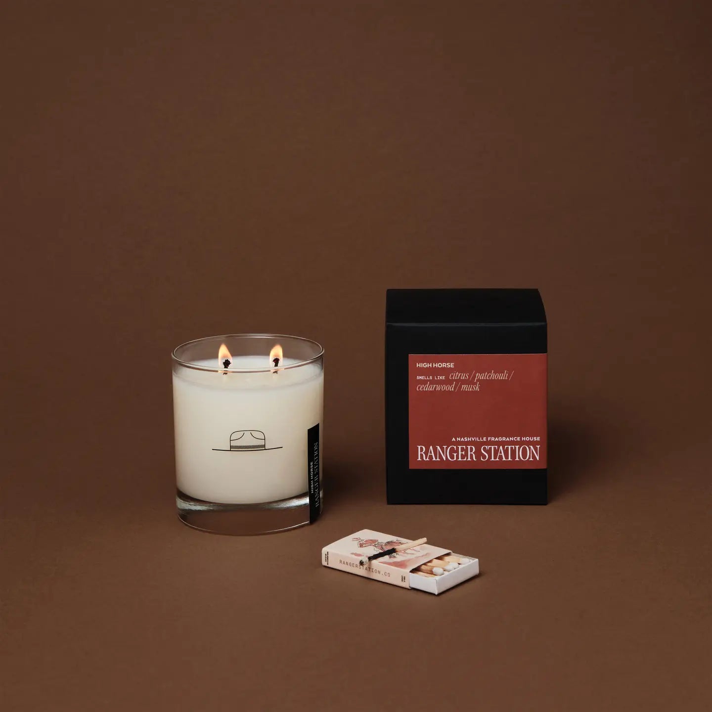 A double-wick Ranger Station Candle made with premium soy wax in a clear glass sits beside a black and red candle box and a matchbox, all arranged on a brown surface with a brown background.
