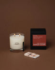 A double-wick Ranger Station Candle made with premium soy wax in a clear glass sits beside a black and red candle box and a matchbox, all arranged on a brown surface with a brown background.