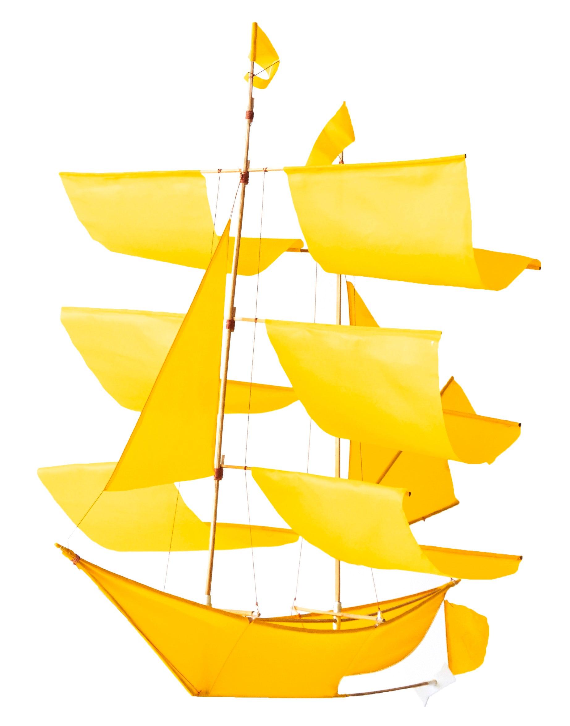The Faire Sailing Ship Kite features multiple bright yellow sails and a brown mast, evoking a handmade aesthetic. Photographed on a white background.