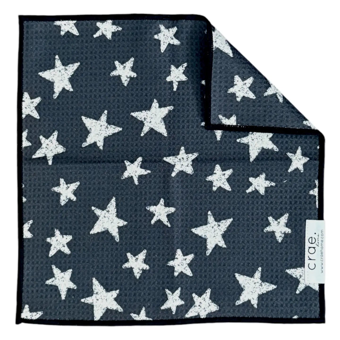 A Faire washcloth in dark gray with a textured feel, white star pattern of varying sizes, and a black border. One corner is folded to reveal the reverse side and a white "crae" tag. Made from eco-friendly materials.