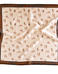 The Faire Wild Rag Silk Scarf features a cream background, brown border, and repeating horses and trees print. Its gently wrinkled silk adds charm, making it a stylish cowgirl accessory.
