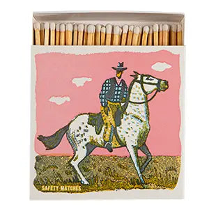 AG Safety Matches by Faire feature a cowboy in a hat, plaid shirt, and yellow pants riding a spotted white horse under a pink sky—ideal as decorative gift drawer accessories.