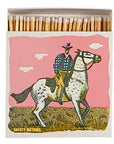 AG Safety Matches by Faire feature a cowboy in a hat, plaid shirt, and yellow pants riding a spotted white horse under a pink sky—ideal as decorative gift drawer accessories.