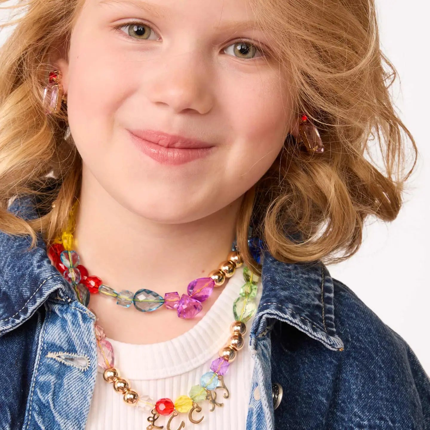 A young child with light hair smiles, wearing necklaces and earrings crafted from the Faire Make It Alphabet DIY Bead Kit. They're dressed in a blue denim jacket over a white top against a plain white background.