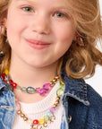 A young child with light hair smiles, wearing necklaces and earrings crafted from the Faire Make It Alphabet DIY Bead Kit. They're dressed in a blue denim jacket over a white top against a plain white background.