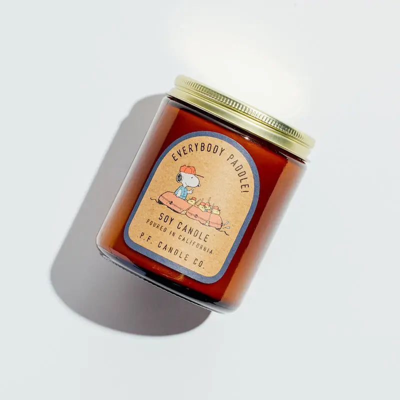 A Limited For Peanuts™– Standard Candle from Faire features a brown apothecary amber jar with a gold lid. It has an illustration of a person paddling with ducks and the text "Soy Wax Candle, Made in California." Fine fragrance oils blend beautifully as shadows are cast beside the jar.