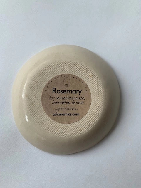 An upside-down Ring Bowl by Faire is shown, revealing its textured base and a brown label: "Rosemary for remembrance, friendship & love. csfceramics.com"—a jewelry catchall inspired by botanical symbolism.
