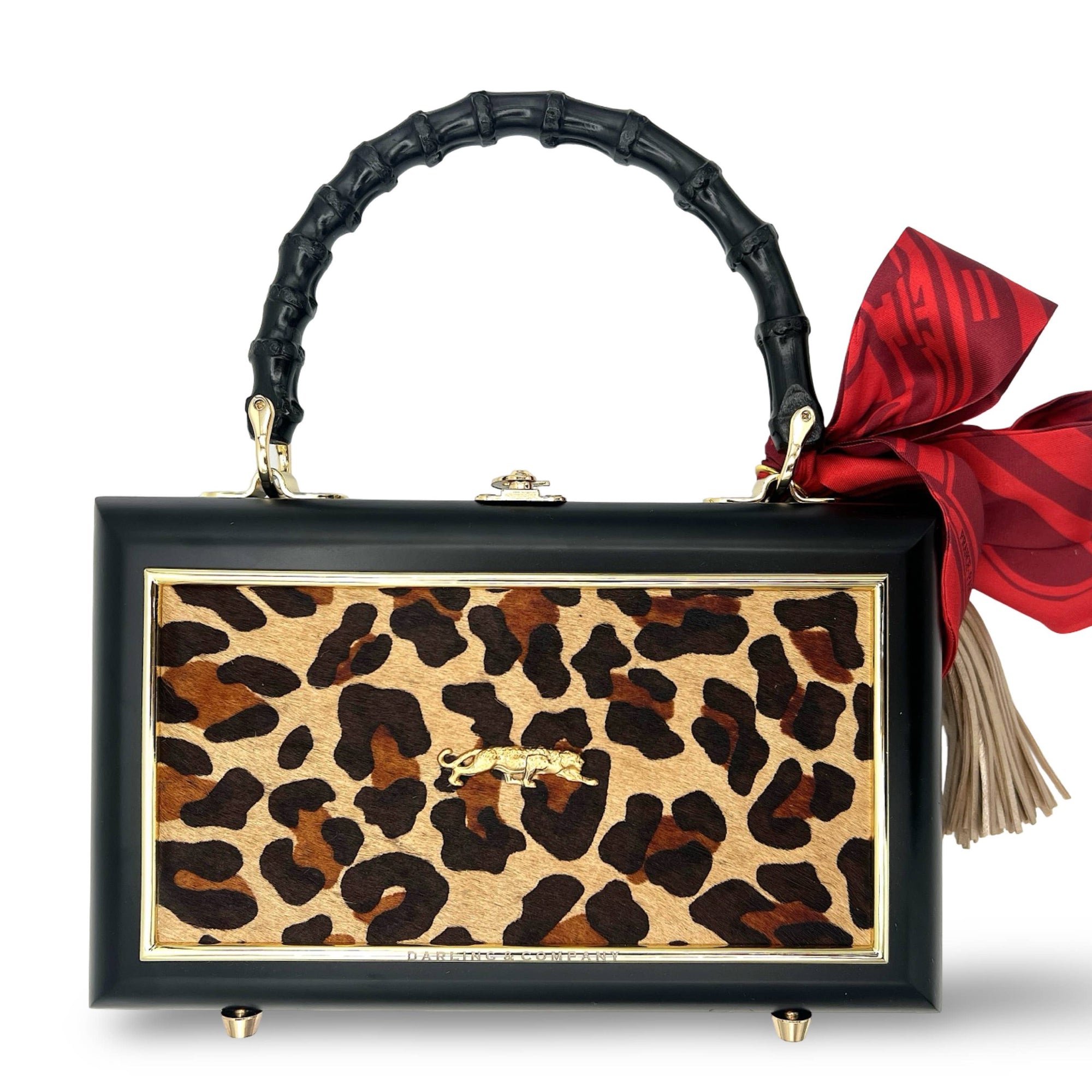 The Faire Leopard Supreme Bag features a rectangular leopard print cowhide design with a black frame, gold accents, and black bamboo handle. It’s finished with a bold red bow, beige tassel, four gold feet, and an eye-catching gold leopard bauble.