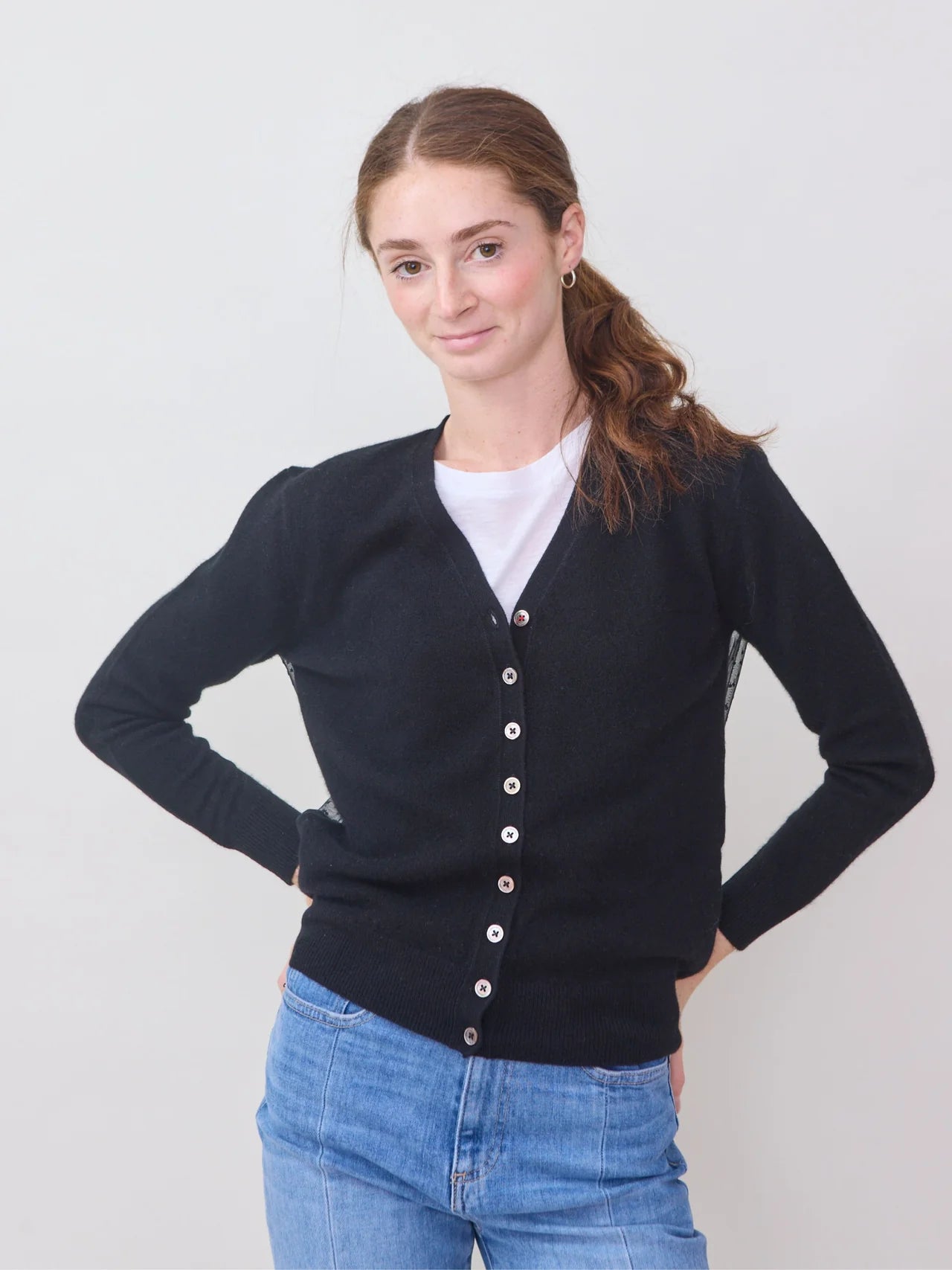 A woman with light skin and long brown hair in a ponytail wears the Brazeau Tricot Tulle Back Cardigan over a white shirt and light blue jeans, standing with hands on her hips and smiling slightly against a plain background.