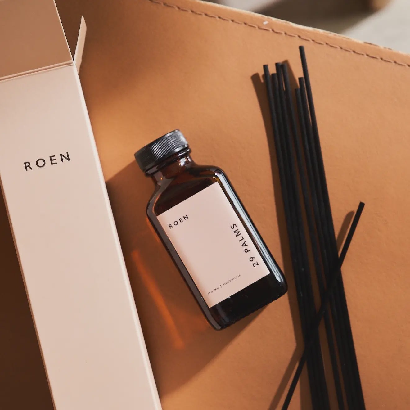 A small amber bottle labeled "29 Palms Reed Diffuser" rests on a tan surface beside black diffuser sticks, exuding hints of eucalyptus. An open box with the brand name "Faire" is on the left, enhancing the minimalist and elegant aesthetic.