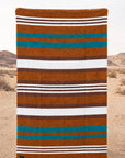 Handwoven Faire Canyon Cove blanket with brown, white, and teal stripes by Bungalow, displayed in desert landscape.