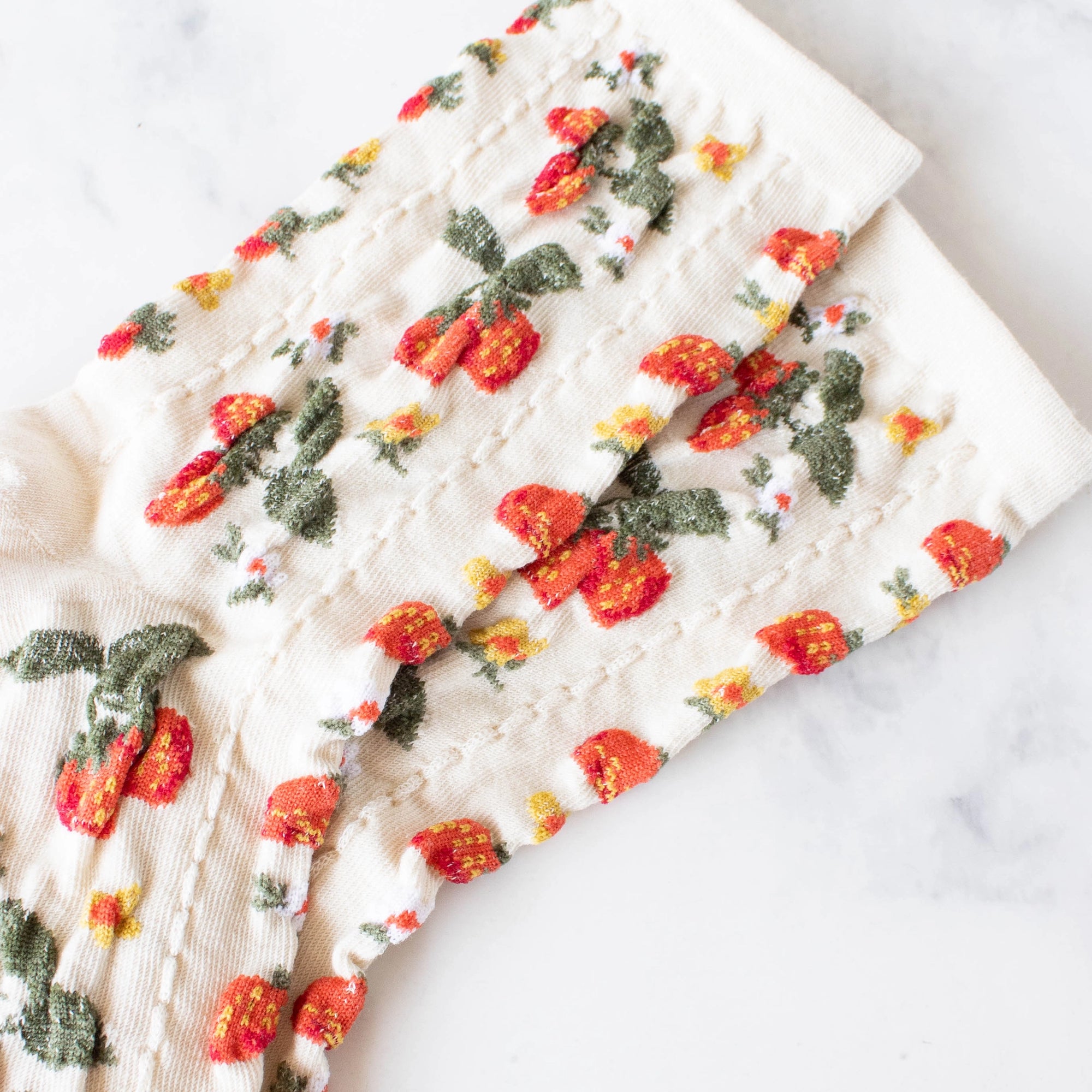 The Faire Strawberry Farm Casual Socks are cream-colored with a textured knit and embroidered red and orange strawberries plus green leaves, laid flat on a white marble surface.