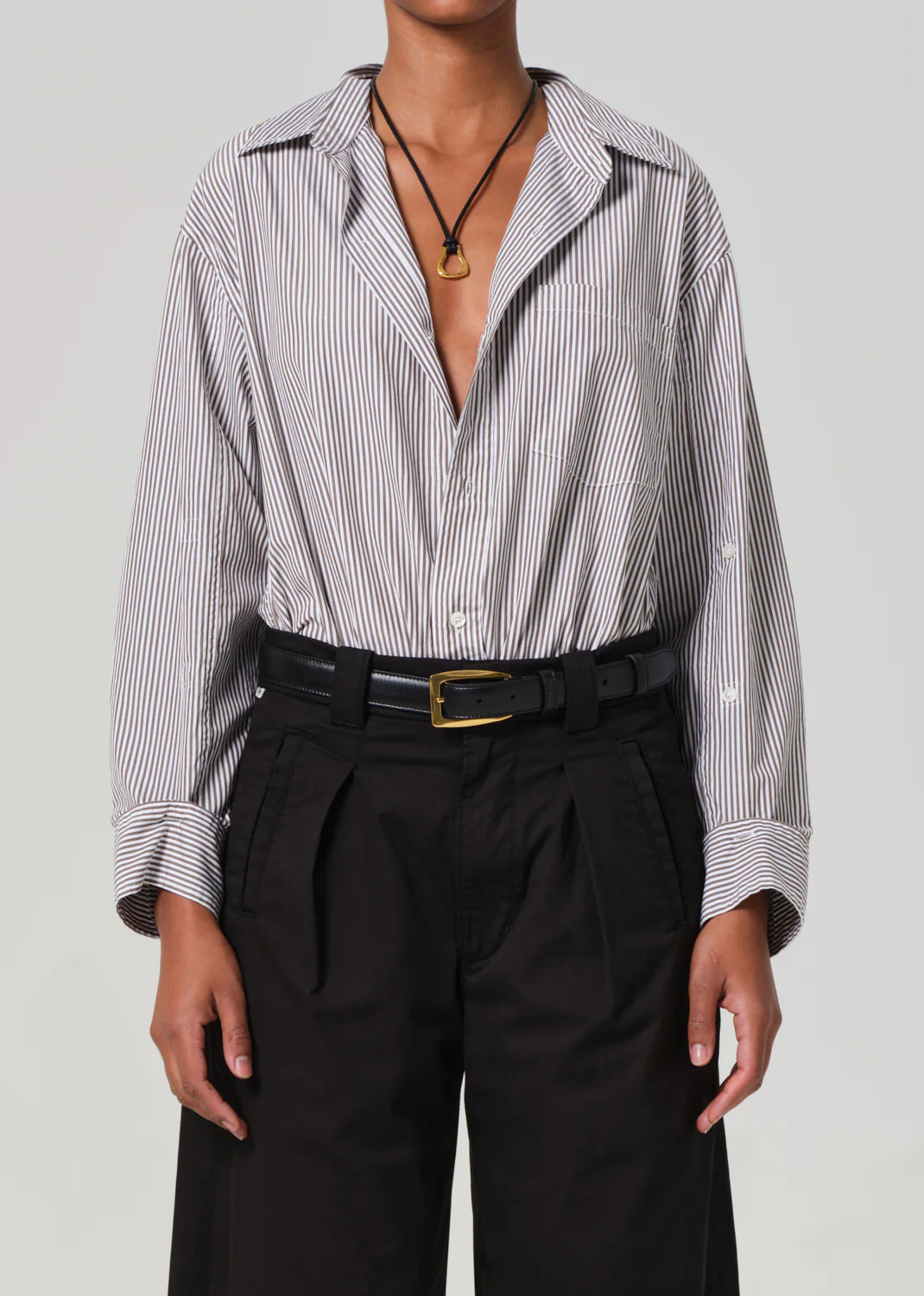Effortless style is achieved with a relaxed, oversized pinstriped Kayla Shirt by Citizens Of Humanity/AGOLDE, showcasing rolled sleeves and a partially unbuttoned look. Pair it with black high-waisted pants, a gold-buckled black belt, and a pendant necklace for chic sophistication.