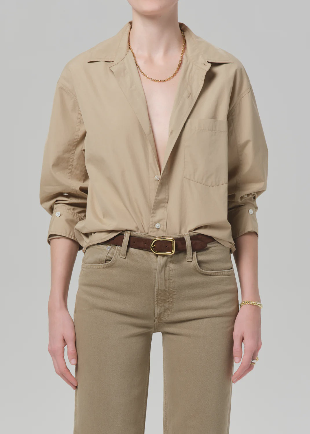 Wearing the Kayla Shirt Catalina by Citizens Of Humanity/AGOLDE, a beige cotton long-sleeve button-up with top buttons undone, tucked into tan pants and styled with a brown belt, gold necklace, and bracelet. Head is not visible.