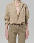 Wearing the Kayla Shirt Catalina by Citizens Of Humanity/AGOLDE, a beige cotton long-sleeve button-up with top buttons undone, tucked into tan pants and styled with a brown belt, gold necklace, and bracelet. Head is not visible.