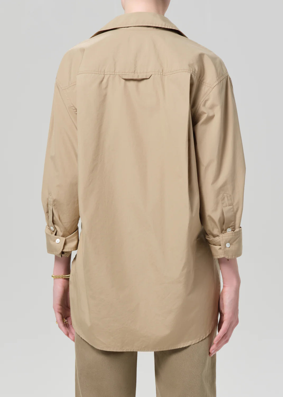 Model shown from the back wearing the Kayla Shirt Catalina by Citizens Of Humanity/AGOLDE—a loose-fitting, beige, long-sleeve poplin shirt with rolled sleeves—paired with beige pants against a plain gray background.