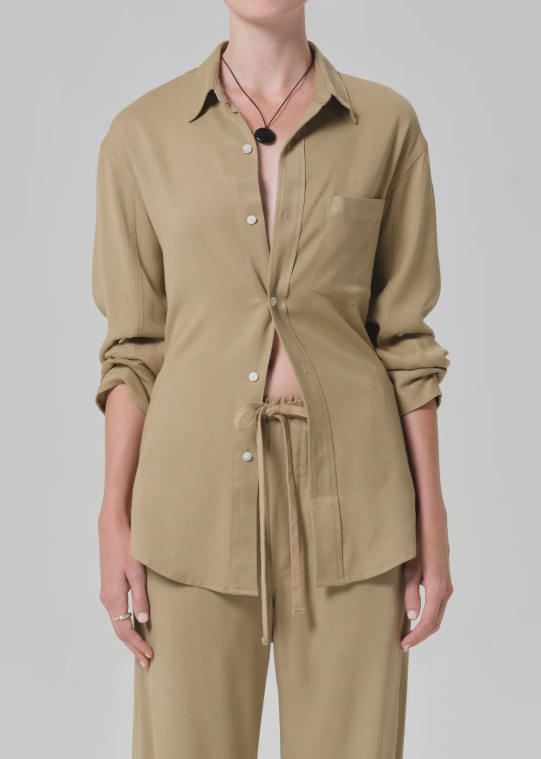 Wearing the kayla shirt in portofino twill by Citizens Of Humanity/AGOLDE, styled with rolled-up sleeves and matching drawstring pants. The partially unbuttoned front is accessorized with a black pendant necklace and rings.