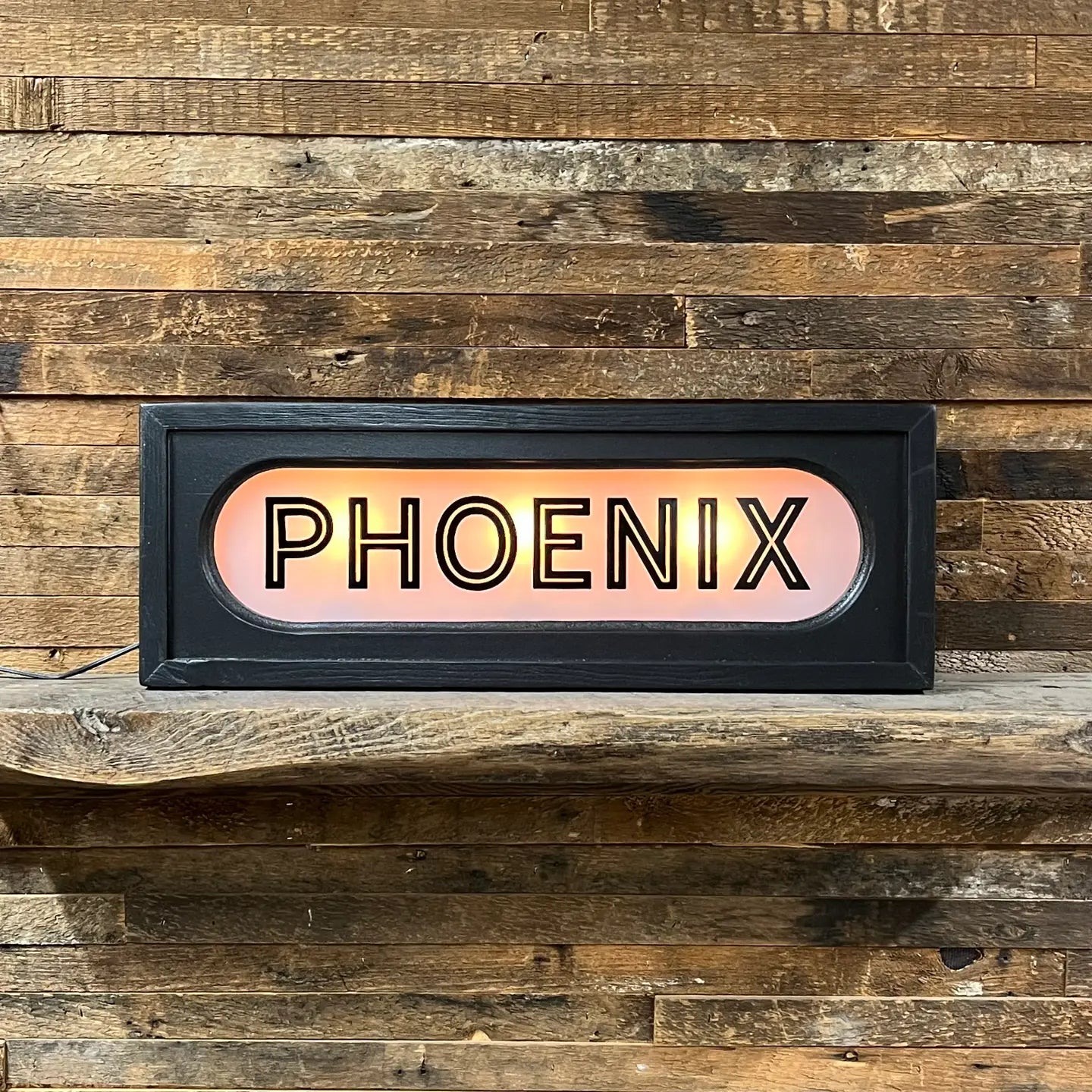 The Faire Lighted Sign, featuring "PHOENIX" in bold black letters, sits on a rustic wooden shelf against a plank wall—a striking personalized decor piece.