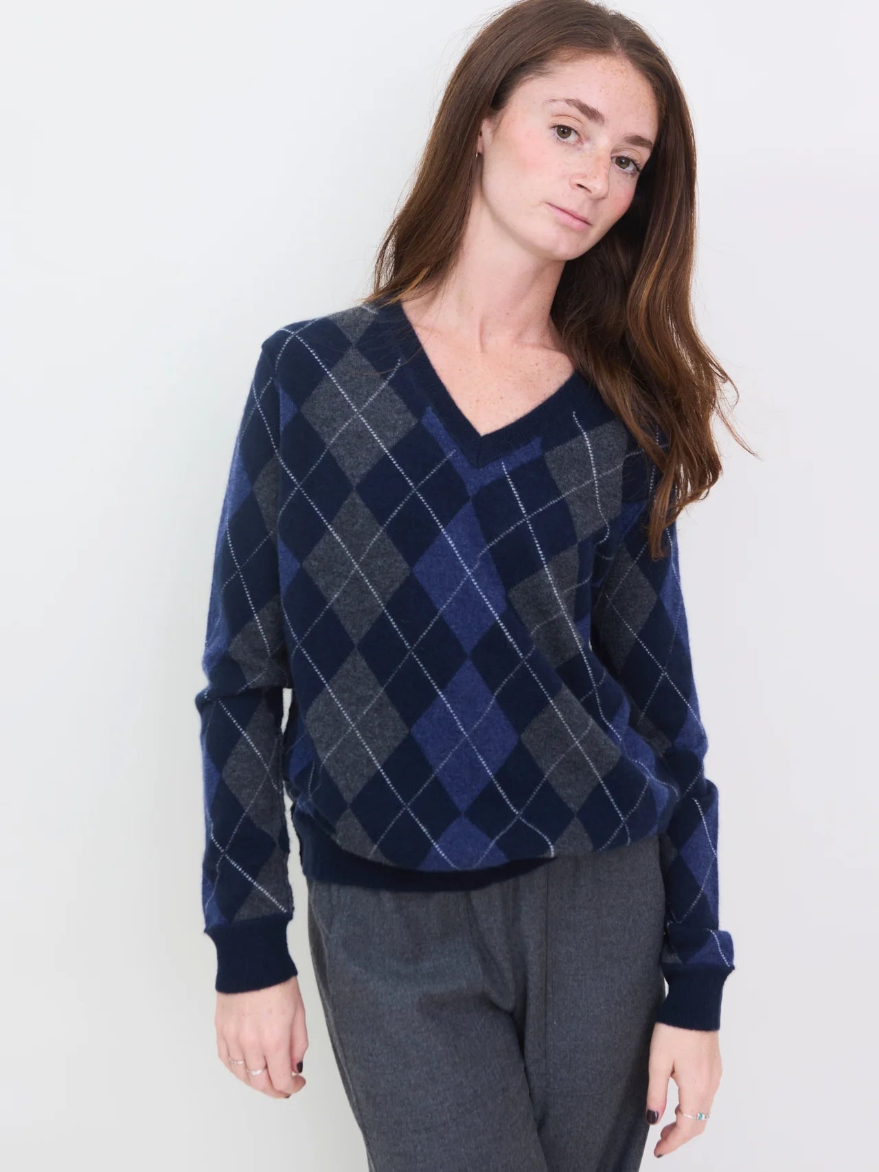 A woman with long brown hair models the Brazeau Tricot Argyle V-Neck in blue and gray, paired with gray pants, standing before a white background and facing the camera with a neutral expression.