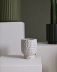 An Adrien Pot by Accent Decor, a textured white ceramic cup, sits on a white pedestal. In the background are green and dark green ribbed ceramic planters with cacti, creating a modern minimalist aesthetic.