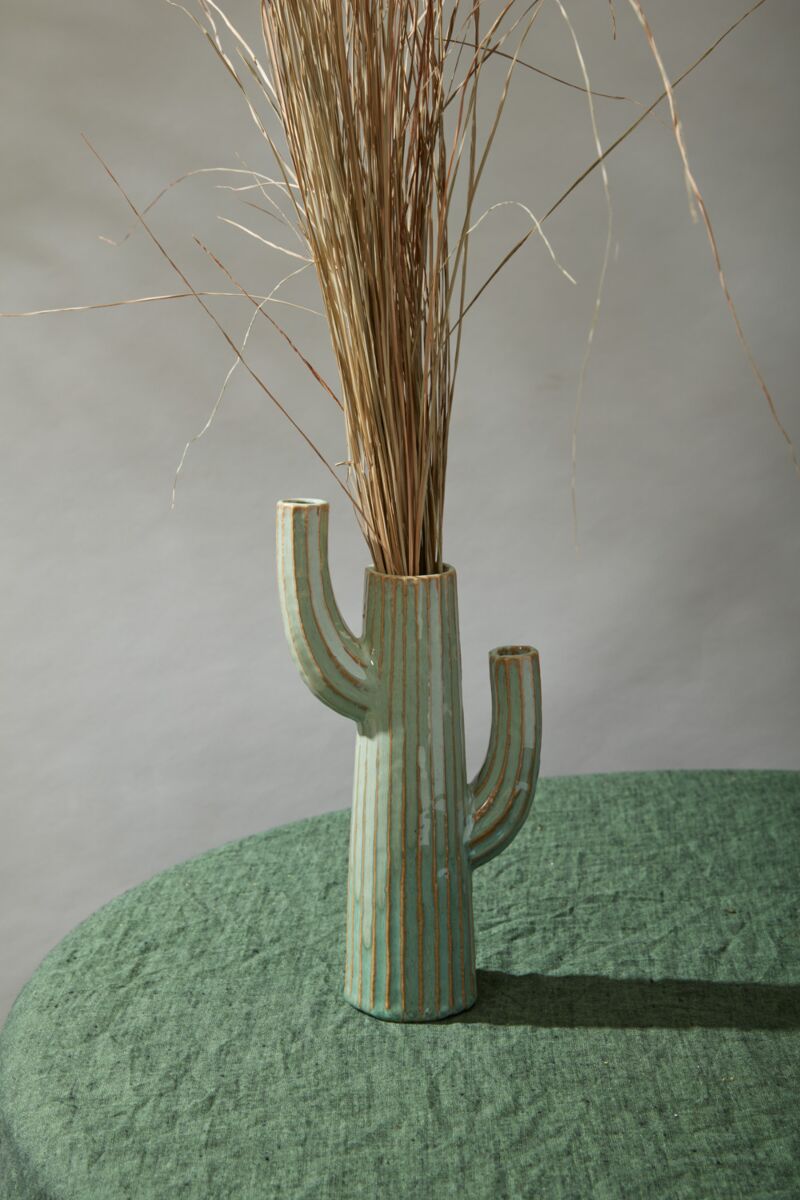 Accent Decor’s Saguaro Vase, a ceramic cactus-shaped piece with green reactive glaze and brown stripes, holds tall dried grass on a textured green surface against a light gray background.
