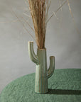 Accent Decor’s Saguaro Vase, a ceramic cactus-shaped piece with green reactive glaze and brown stripes, holds tall dried grass on a textured green surface against a light gray background.