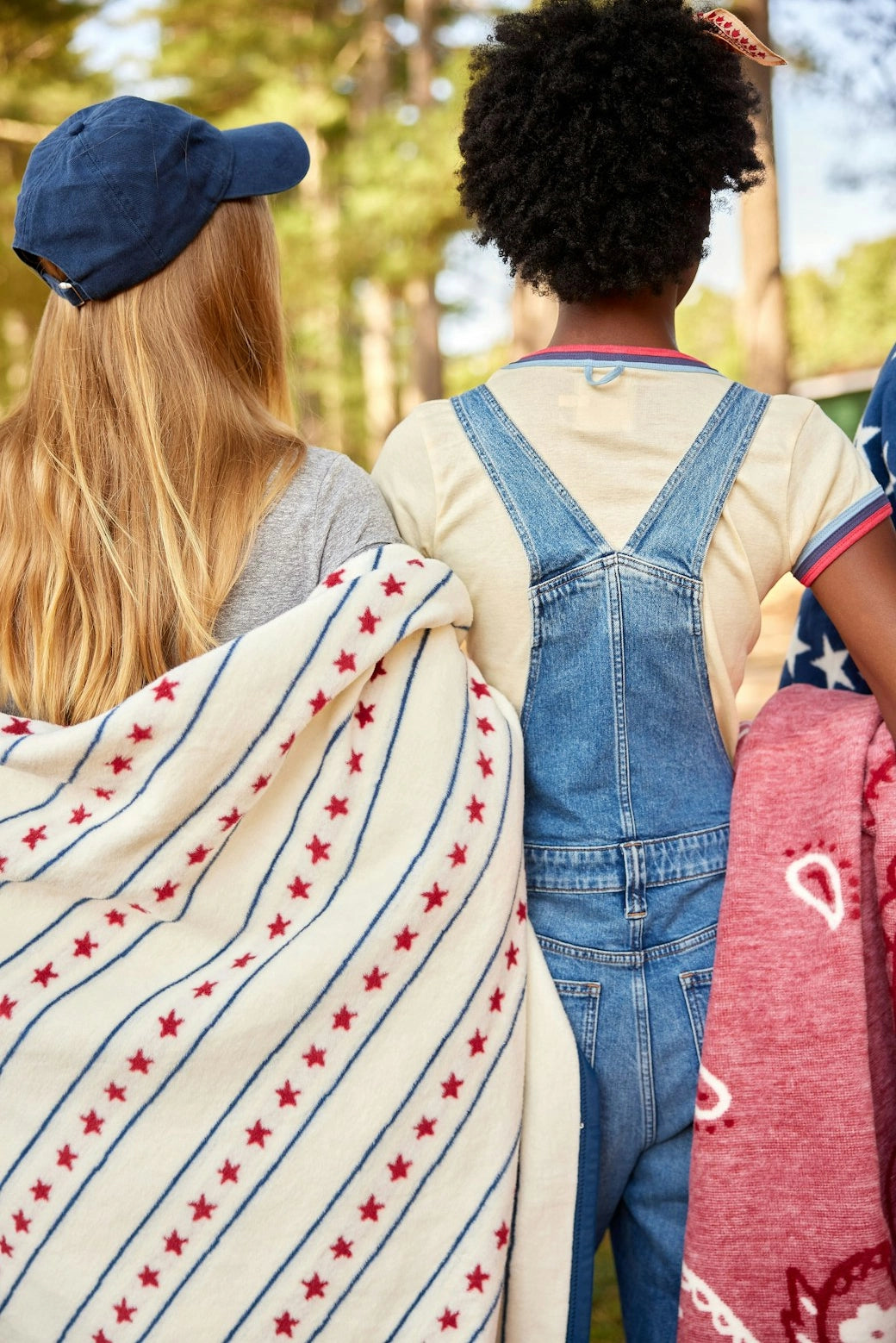 Two children stand outdoors with backs turned. One, in a blue cap, drapes the Faire Stars & Stripes Blanket; the other, wearing denim overalls and a cream shirt, holds a red blanket.