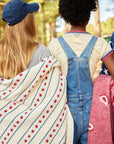 Two children stand outdoors with backs turned. One, in a blue cap, drapes the Faire Stars & Stripes Blanket; the other, wearing denim overalls and a cream shirt, holds a red blanket.