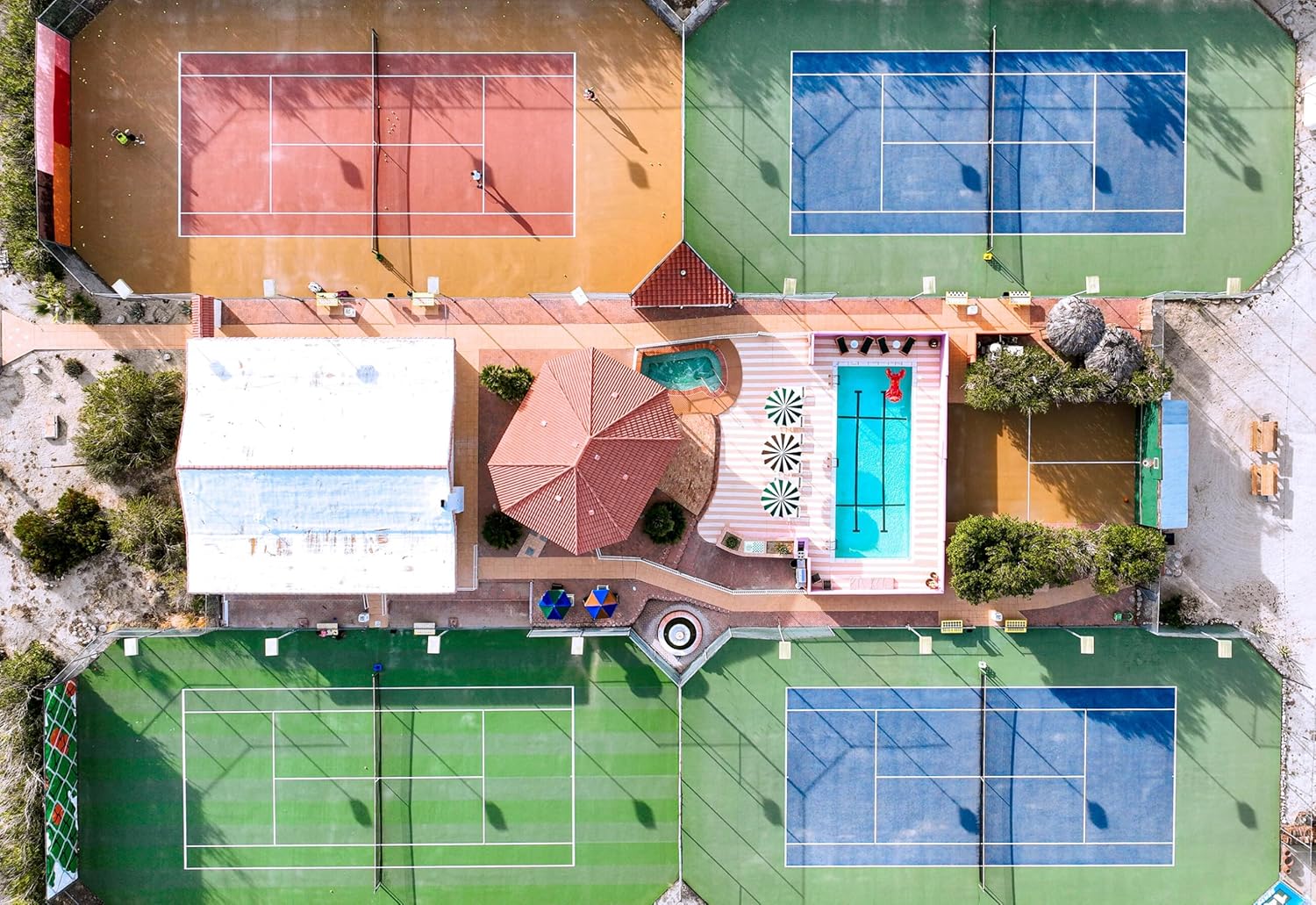 The Faire Tennis Court photo by Nick Pachelli showcases an aerial view of four vibrant courts, a central building, swimming pool, hot tub, lush landscaping, and two people playing tennis.