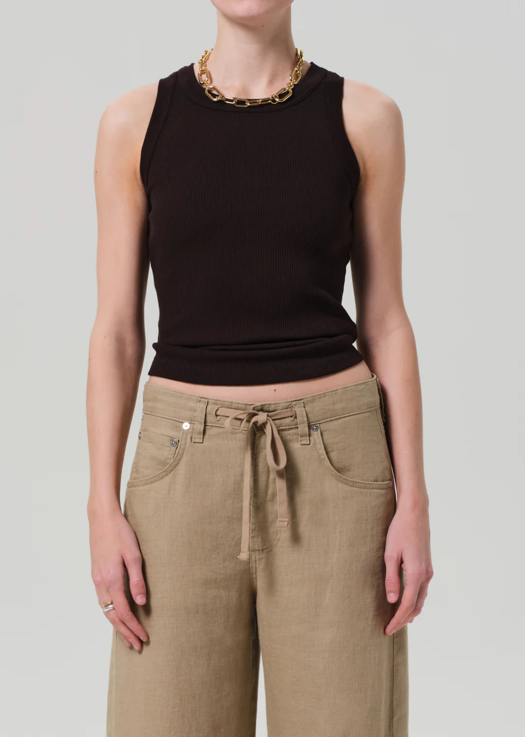 Wearing the Isabel Rib Tank by Citizens Of Humanity/AGOLDE in black, paired with tan wide-leg pants with a tie waist, a gold chain necklace, and rings—an ideal layering piece for versatile styling against a light gray background.