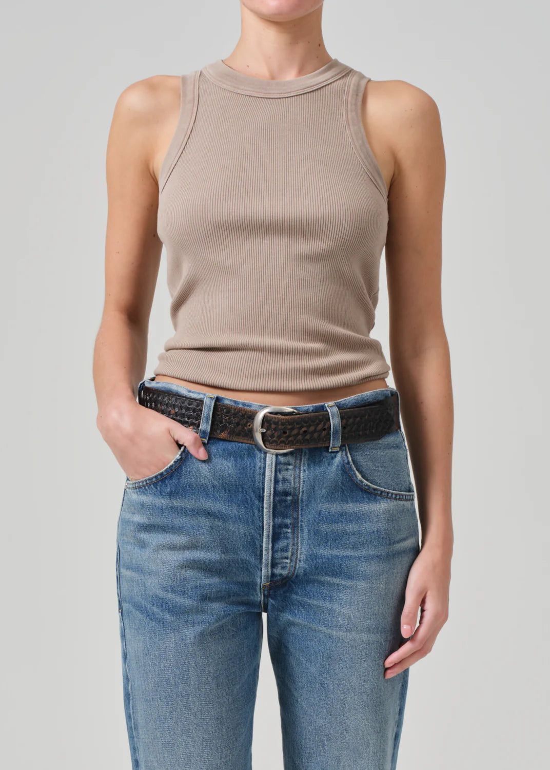 A person stands against a plain background, wearing the Isabel Rib Tank by Citizens Of Humanity/AGOLDE in beige, made from organic cotton, paired with blue jeans and a black woven belt. With one hand casually tucked in their pocket, they effortlessly showcase this versatile layering piece.